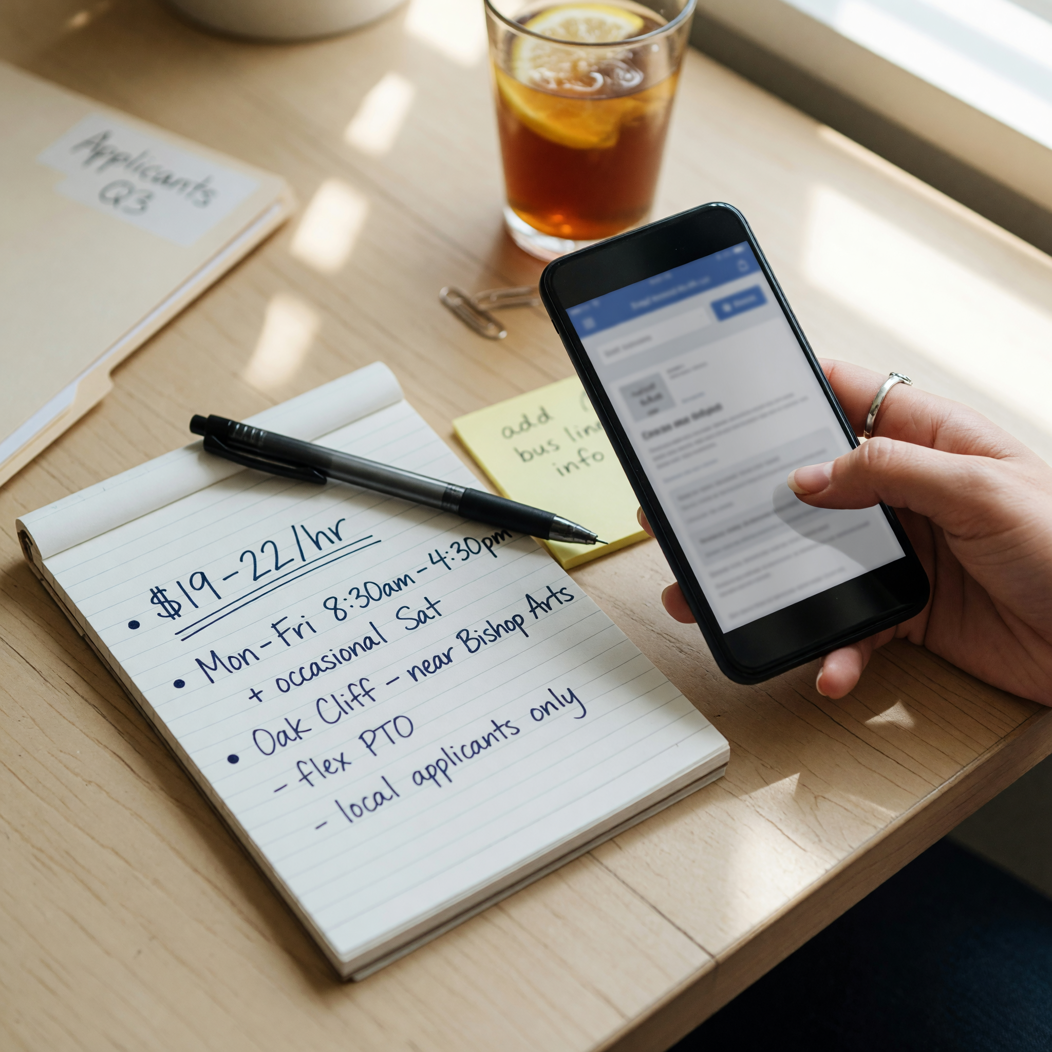 A hiring manager drafting a clear Dallas job post on a phone beside a notepad listing pay, hours, and neighborhood location