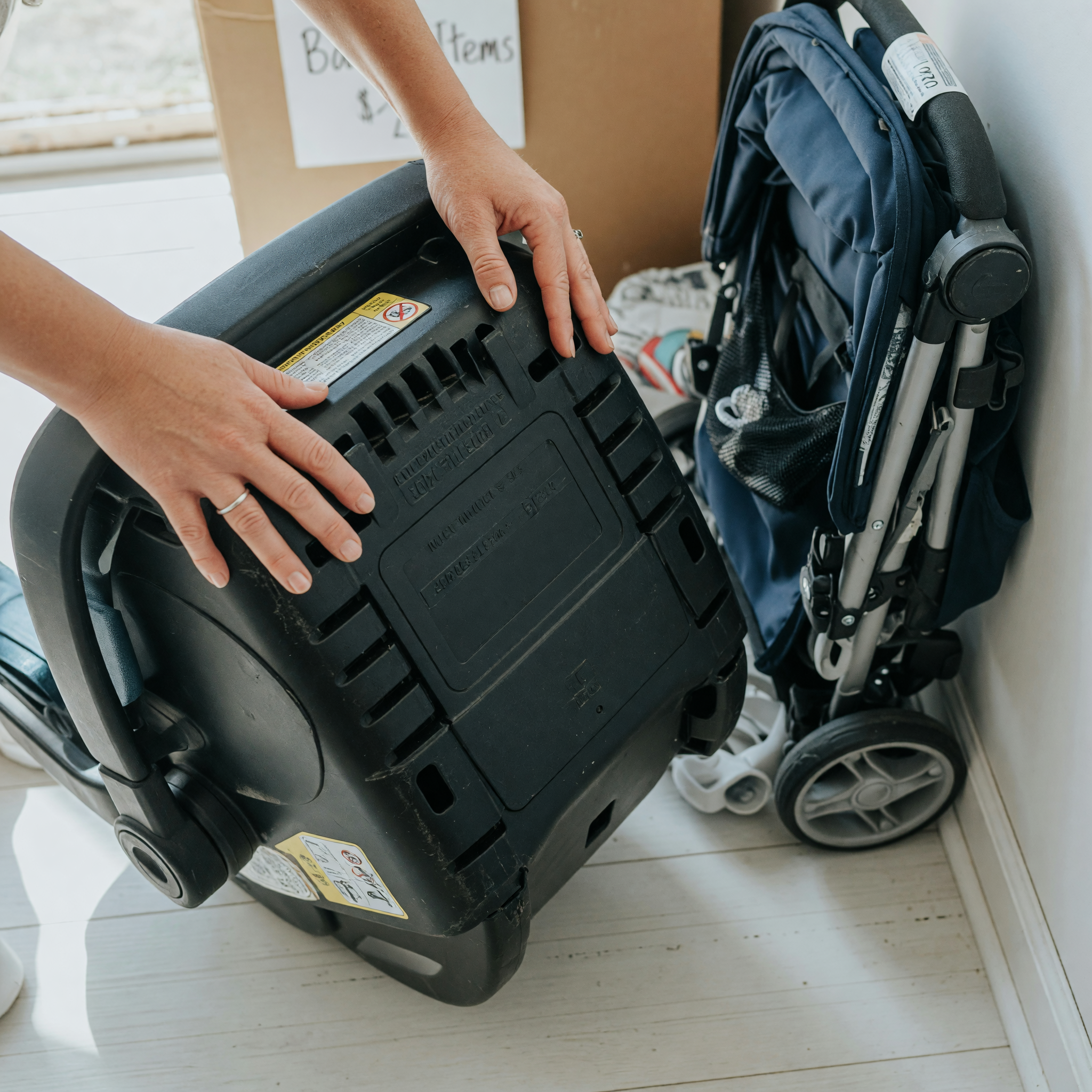 Hands turning over an infant car seat to read the molded manufacture date and expiration label before listing it for sale, with a folded stroller beside it