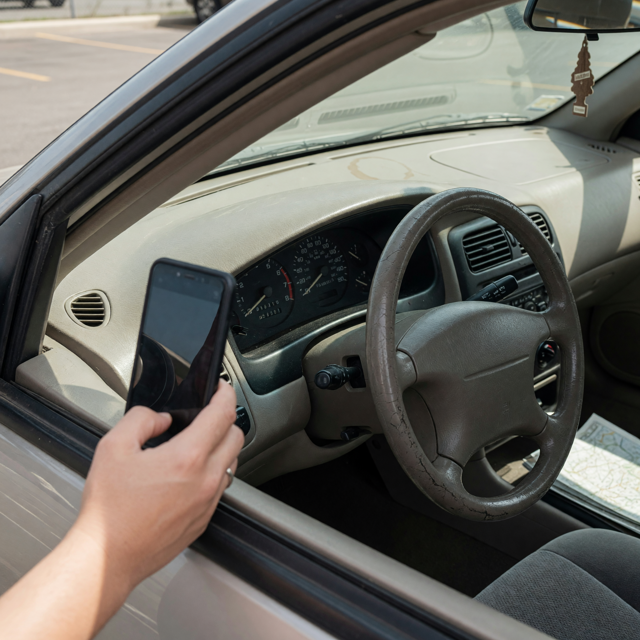 A hand holding a smartphone photographing a used car's odometer and dashboard through the driver window in daylight