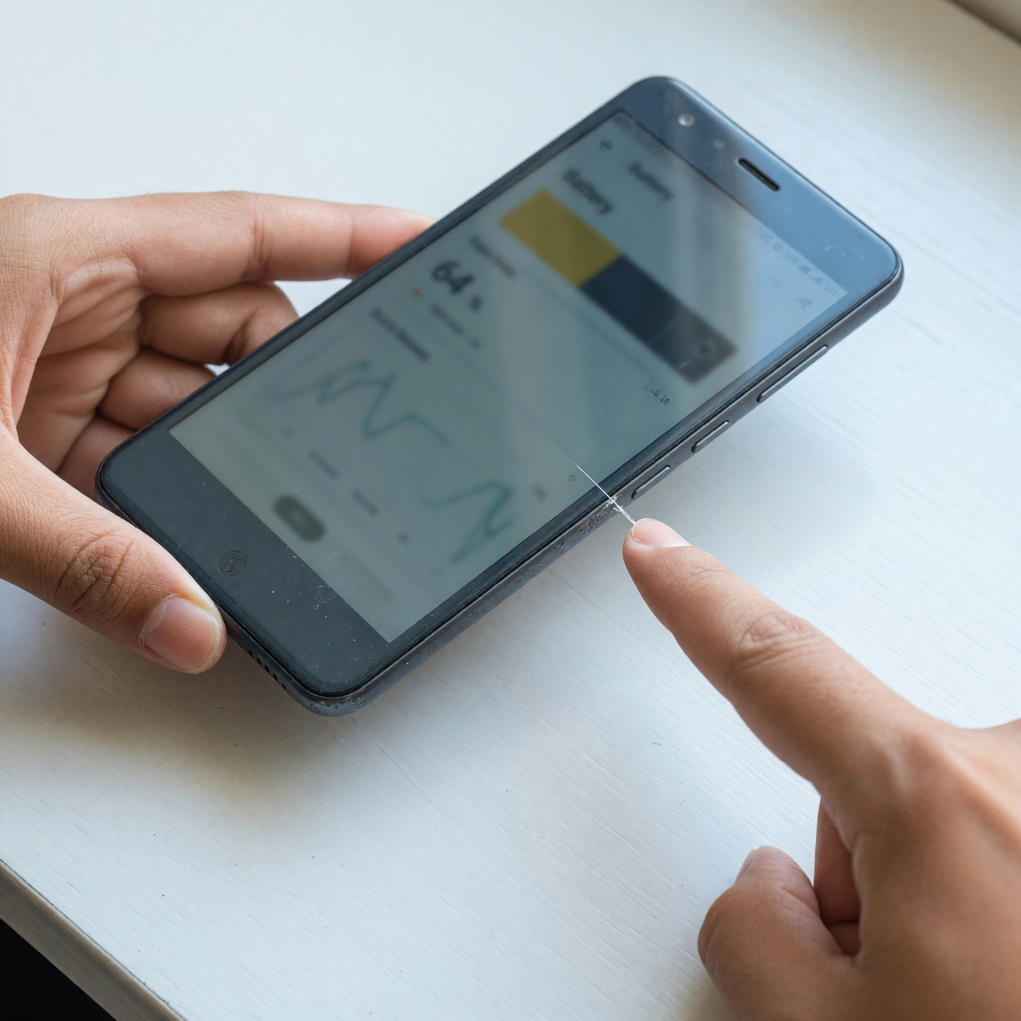 Close-up of hands inspecting a used smartphone, one finger pointing to a small scratch near the screen edge while the settings screen is visible